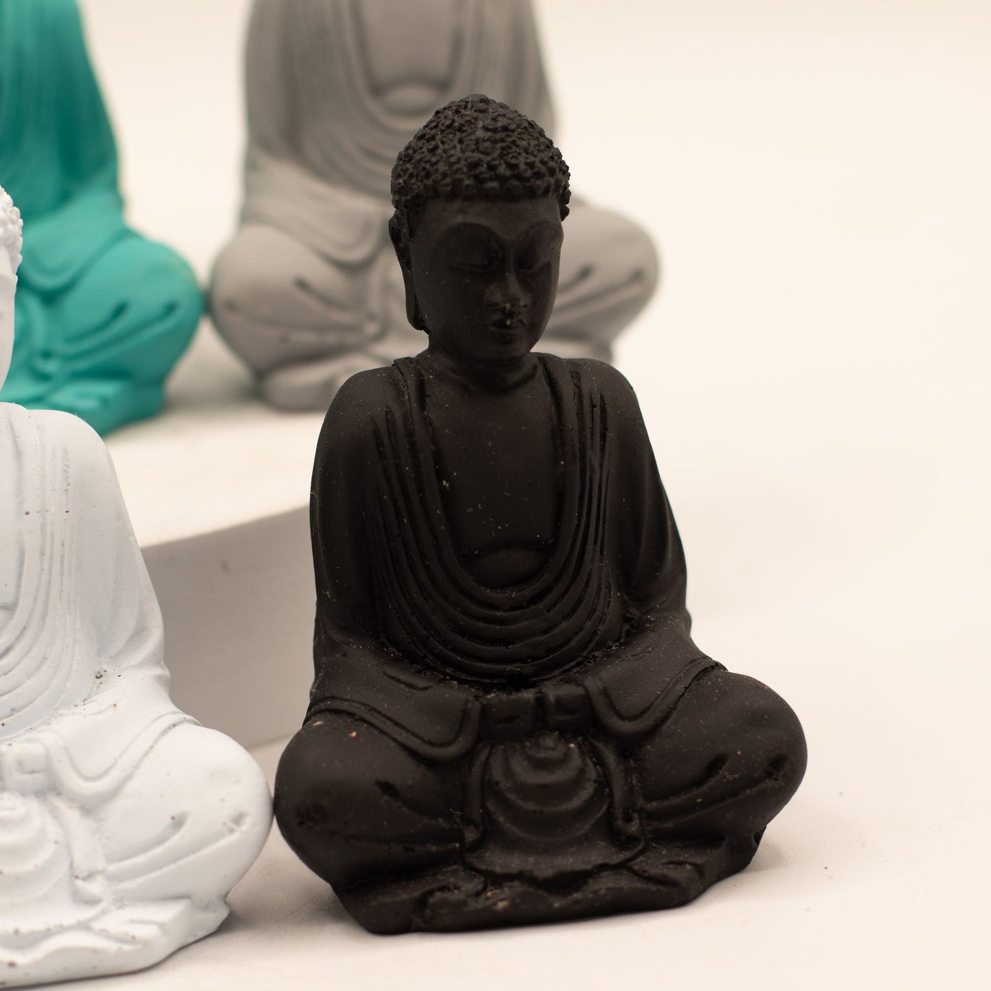 A small black Buddha statue seated in a meditative pose, shown in sharp focus against a softly blurred background with other statue colors partially visible.