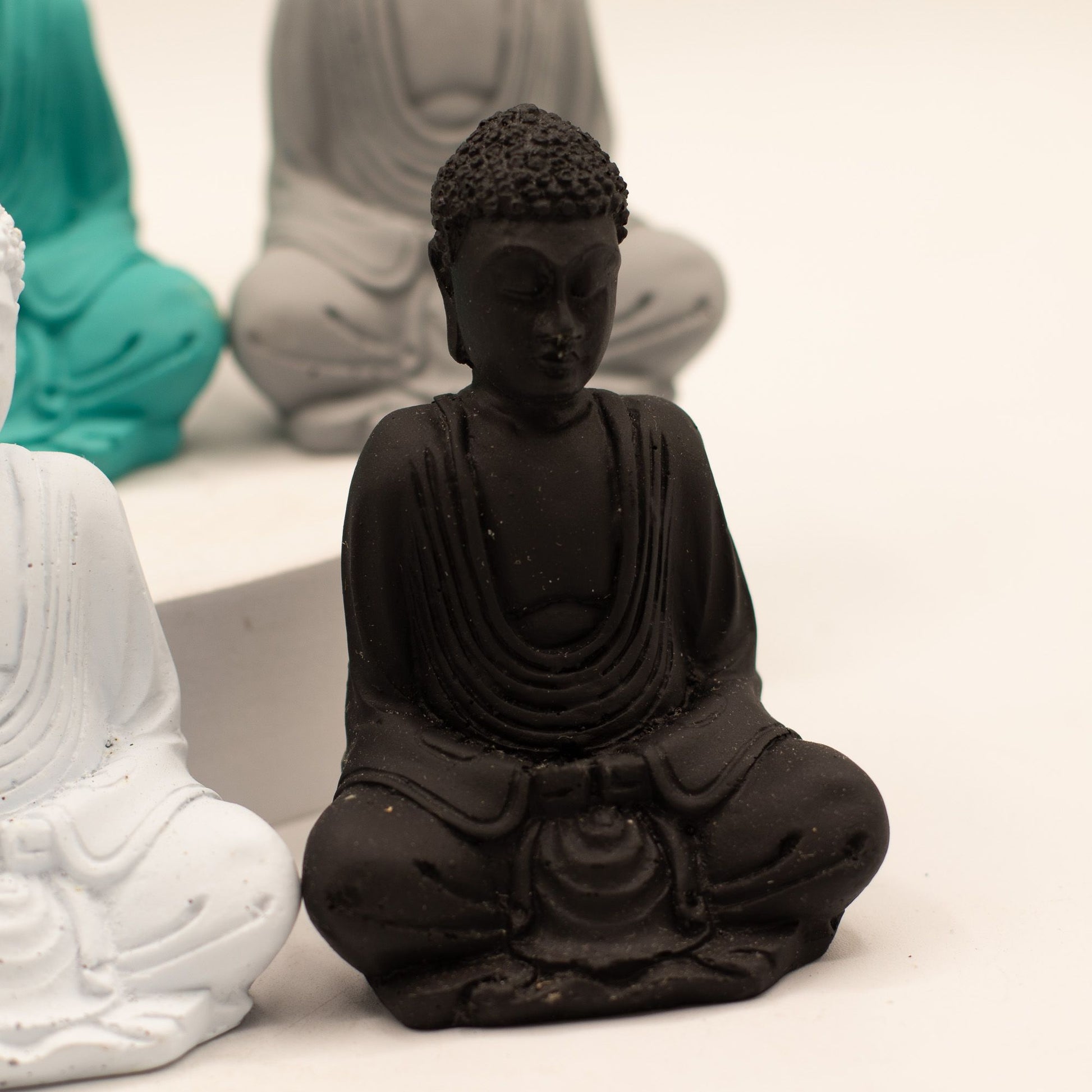 A small black Buddha statue seated in a meditative pose, shown in sharp focus against a softly blurred background with other statue colors partially visible.